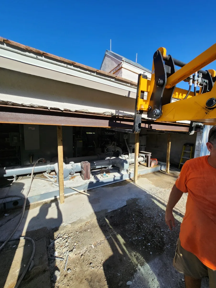 A crane boom positioned at the Naples Heritage pump house wall opening with a steel support beam spanning the gap as the new irrigation station is moved into place.