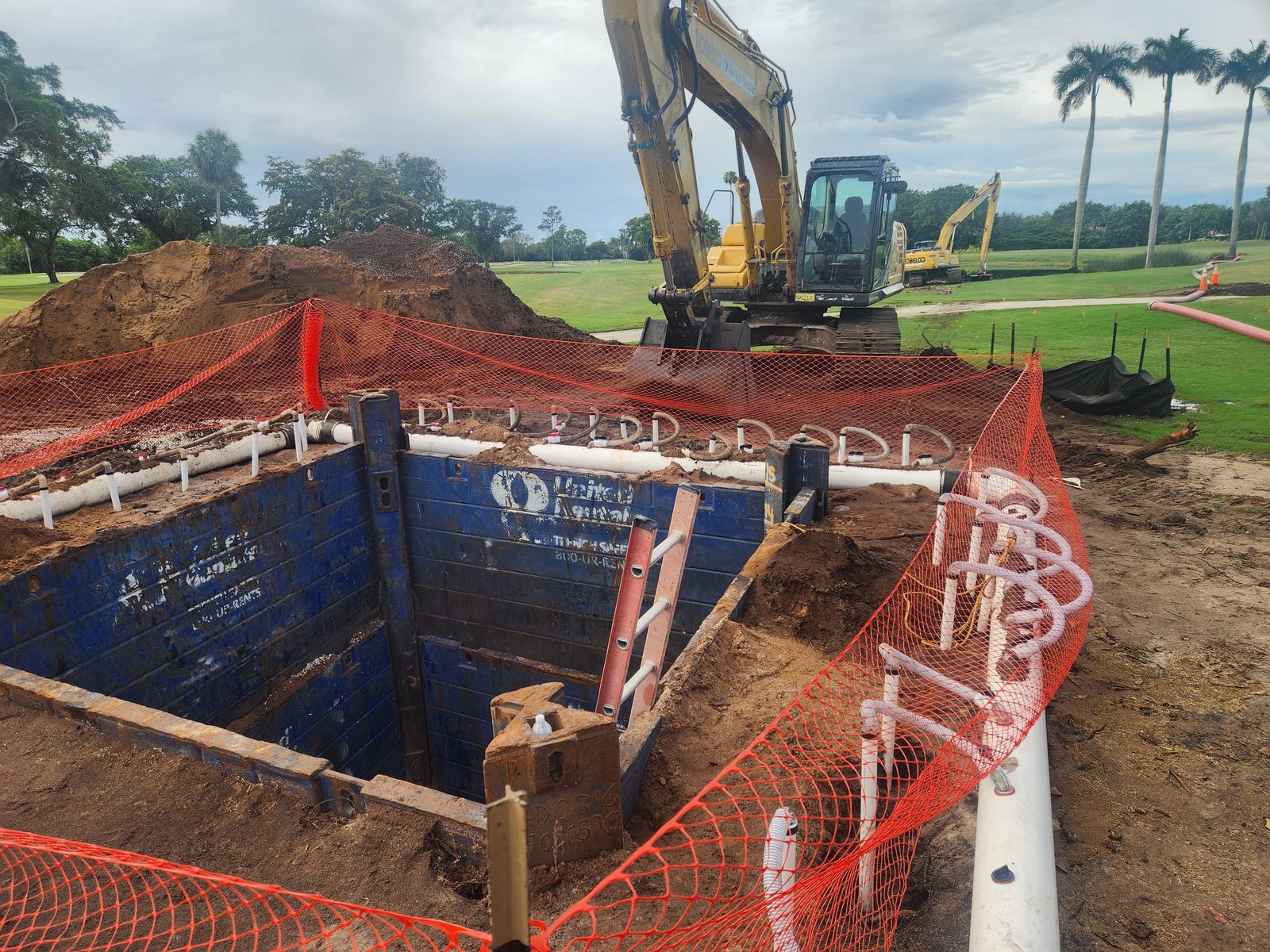 Excavation of the new wet well at Pine Tree Golf Club, with trench-box shoring in place and irrigation mains being routed alongside an excavator on the course