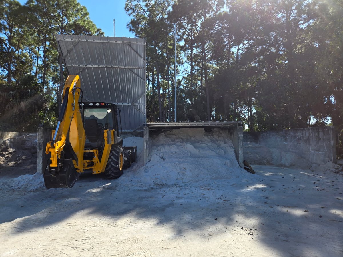 Backhoe accessing sand under a raised soil bin cover on a concrete block storage bin