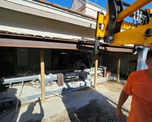 A crane boom positioned at the Naples Heritage pump house wall opening with a steel support beam spanning the gap as the new irrigation station is moved into place.
