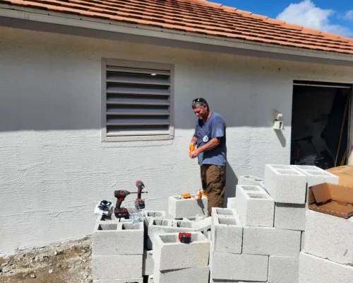 Fresh cinder block staged at the Naples Heritage pump house as the removed wall section is rebuilt beside the louvered vent and finished wall.