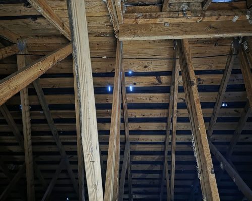 Interior view up into the Old Marsh pump house roof before repair, showing aged wood trusses and deteriorated sheathing with daylight visible through gaps in the boards