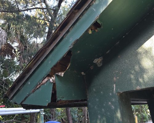 Damaged eave on the Old Marsh pump house before repair, with rotted, broken soffit and fascia crumbling away at the roof edge