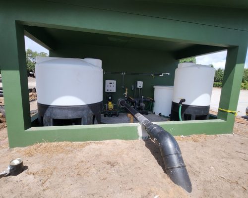 Interior of the completed Pine Tree pumphouse acid-injection bay, with two storage tanks set in a curbed containment area and chemical dosing equipment plumbed to the irrigation main